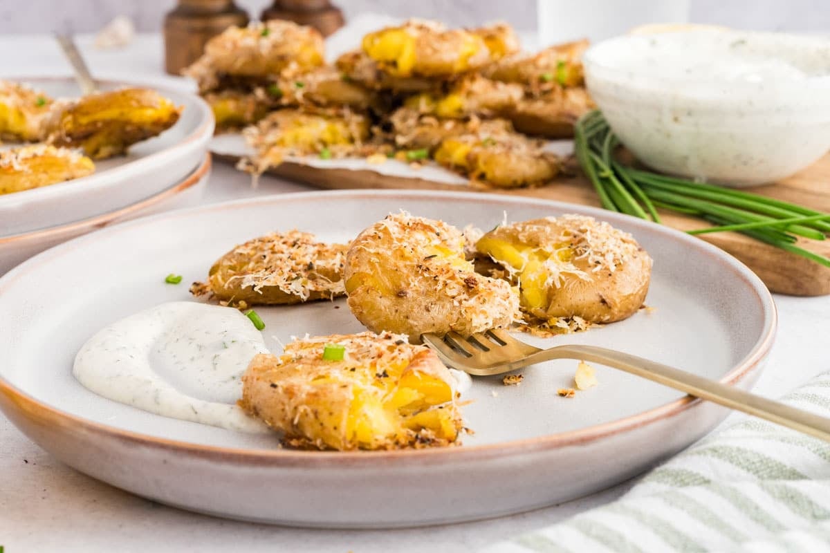 A plate of smashed roasted potatoes with grated cheese, served with a side of creamy dipping sauce and garnished with chives.