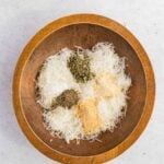 Wooden bowl containing shredded cheese, dried oregano, ground black pepper, garlic powder, and onion powder on a light surface.