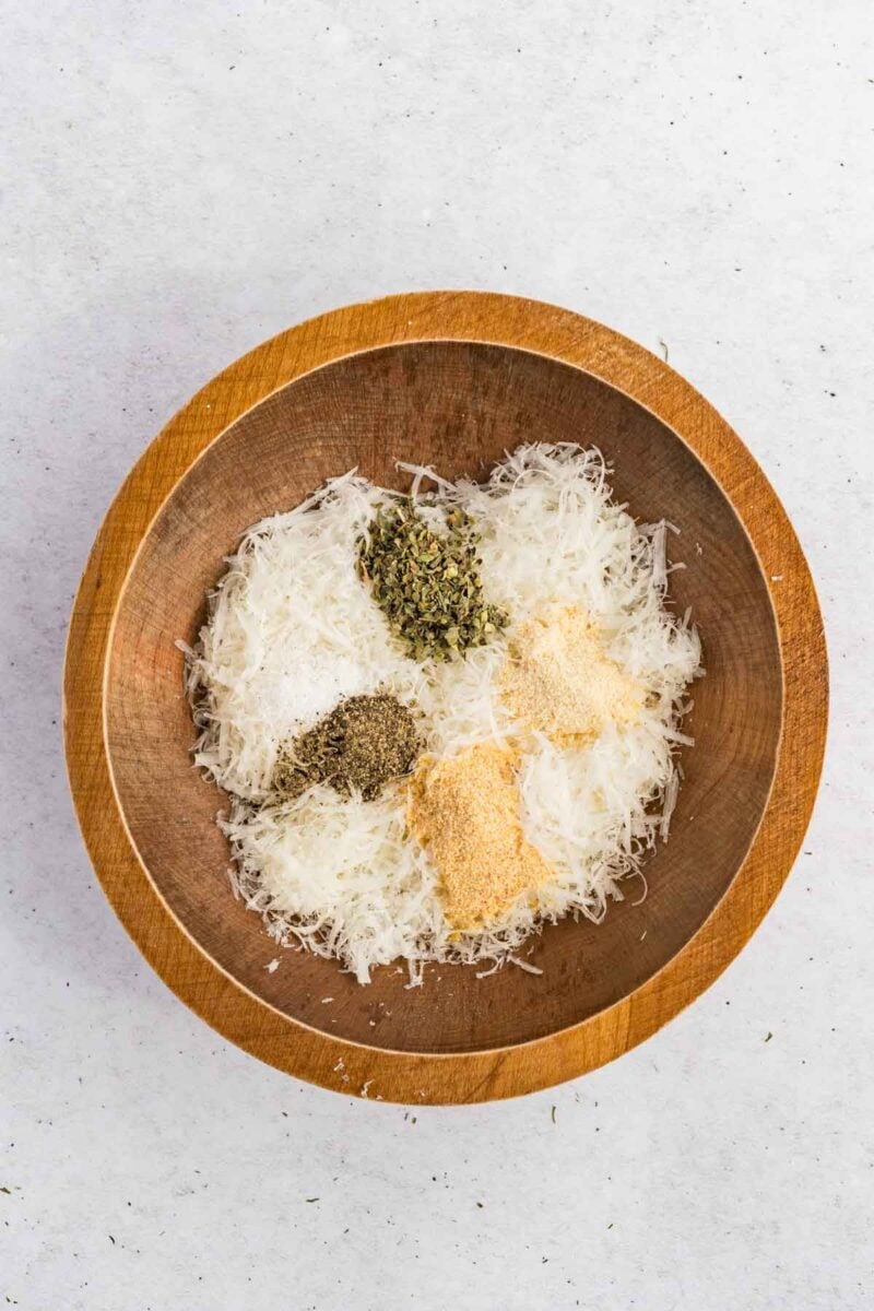 Wooden bowl containing shredded cheese, dried oregano, ground black pepper, garlic powder, and onion powder on a light surface.