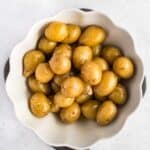 A white scalloped bowl filled with whole baby potatoes, coated with a light glaze, set on a light gray background.