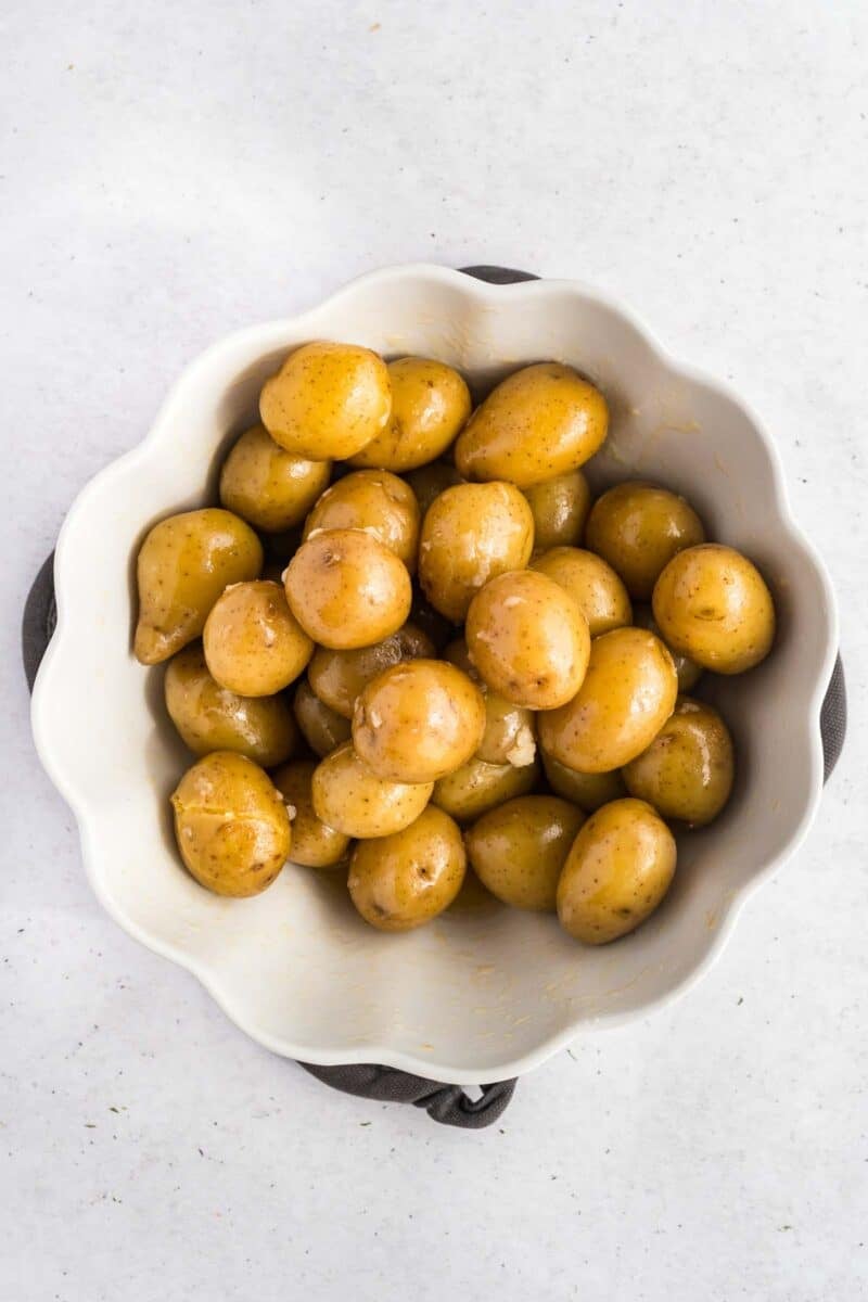 A white scalloped bowl filled with whole baby potatoes, coated with a light glaze, set on a light gray background.