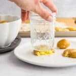 A hand uses a glass jar to press and flatten a boiled potato on a white plate; more potatoes and a bowl are nearby.