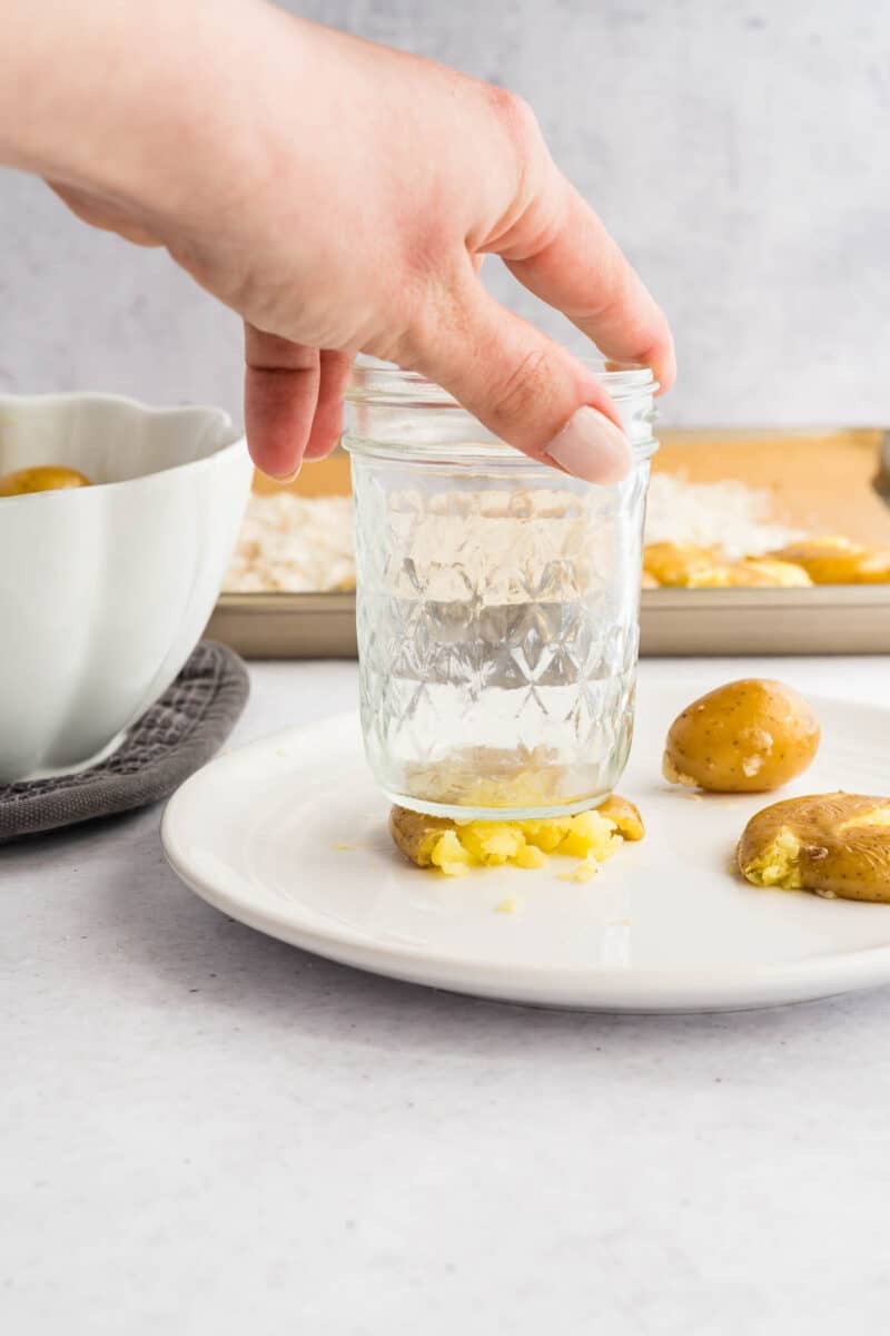 A hand uses a glass jar to press and flatten a boiled potato on a white plate; more potatoes and a bowl are nearby.