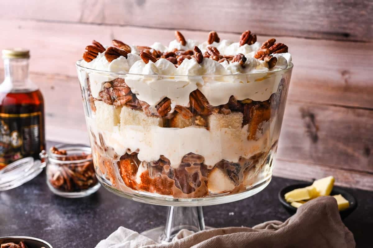 A pecan pie trifle dessert in a glass bowl, featuring layers of cake, cream, pecans, and whipped topping, sits on a dark surface with pecans and syrup nearby.