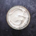 A glass bowl filled with smooth, white whipped cream sits on a dark textured surface, viewed from above.