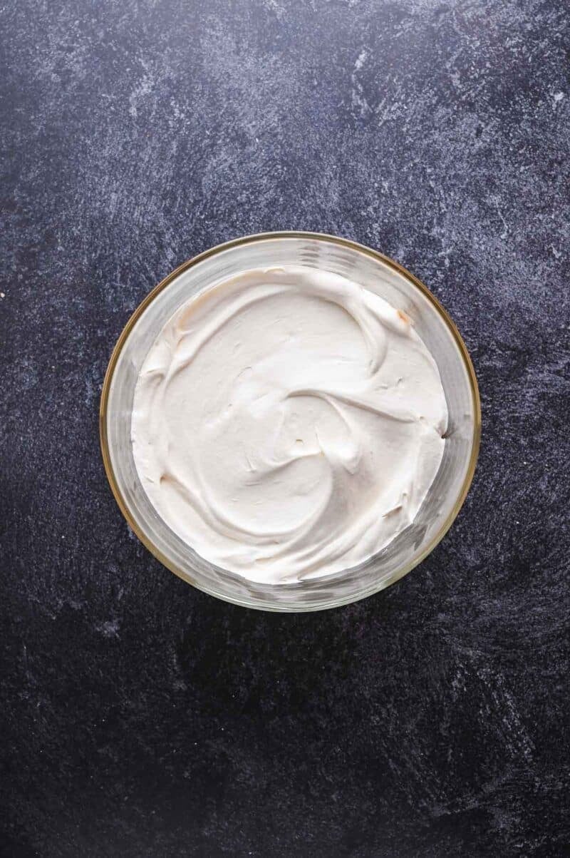 A glass bowl filled with smooth, white whipped cream sits on a dark textured surface, viewed from above.