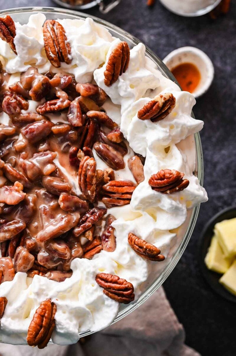 A pecan pie trifle topped with whipped cream and whole pecans, with a center layer of pecans in a syrupy sauce, served in a glass dish.