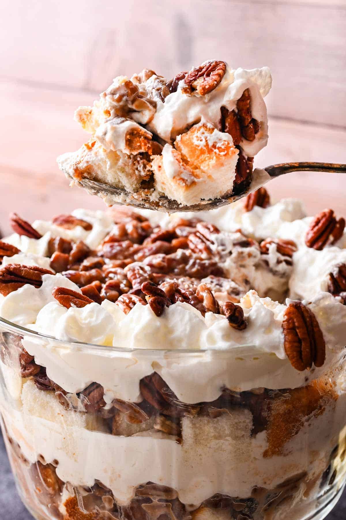 A close-up of pecan pie trifle in a glass bowl with whipped cream, cake pieces, and pecans, with a spoon lifting a serving.