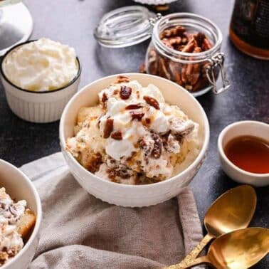 A bowl of pecan pie trifle sits on a table with bowls of pecans, syrup, and whipped cream, plus two gold spoons nearby.