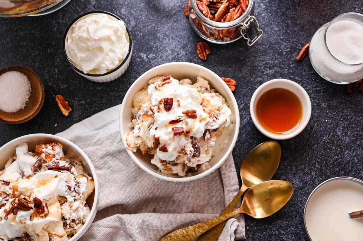 Two bowls of pecan pie trifle on a dark surface, surrounded by bowls of sugar, pecans, vanilla extract, and a jar of cream, with two gold spoons nearby.