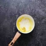 A small saucepan with a wooden handle containing melted butter and cream, placed on a dark textured surface.