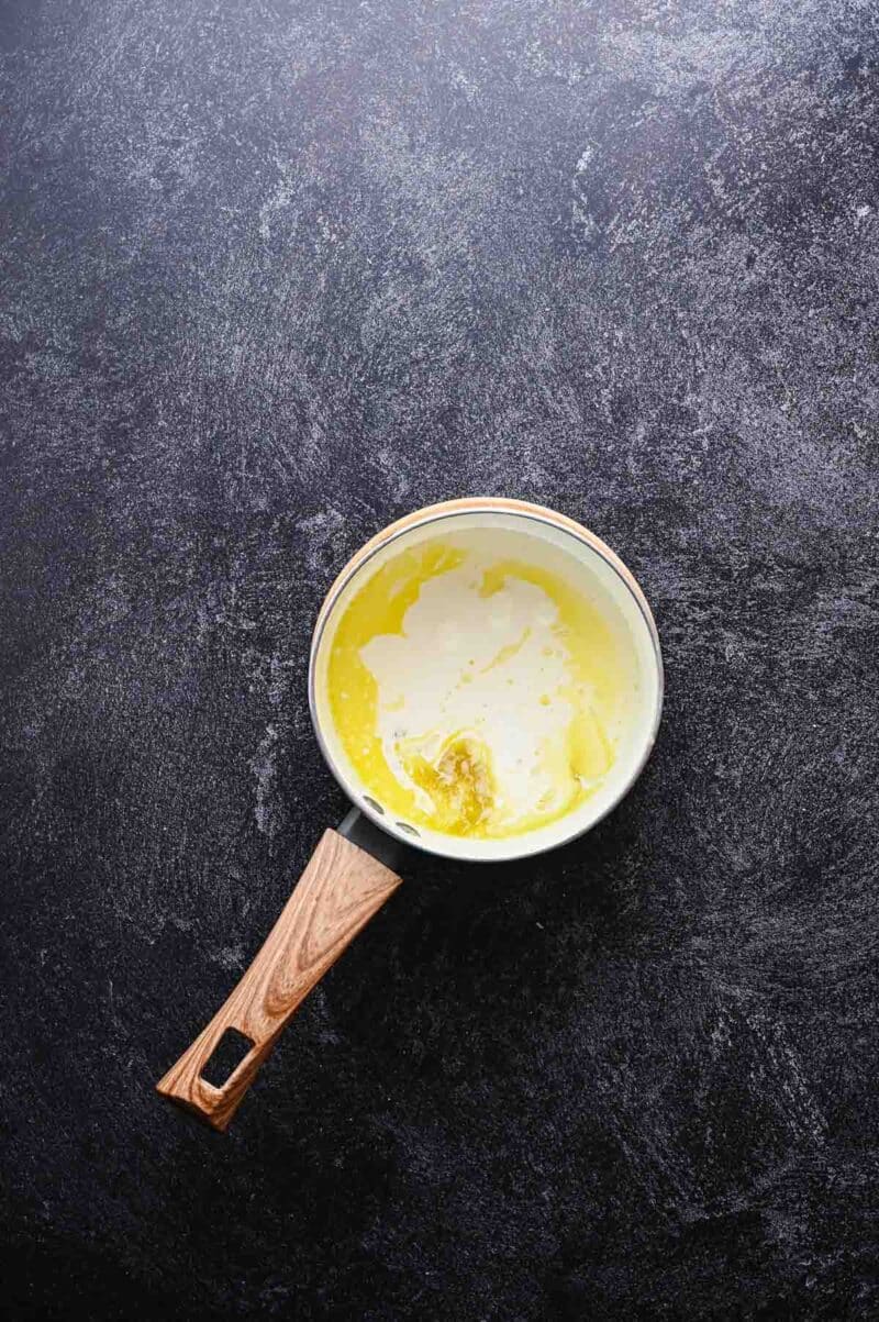 A small saucepan with a wooden handle containing melted butter and cream, placed on a dark textured surface.