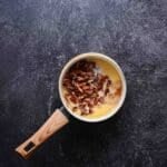 A small saucepan with melted butter and chopped pecans sits on a dark textured surface.