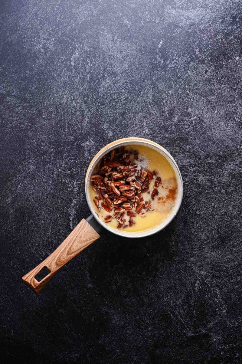 A small saucepan with melted butter and chopped pecans sits on a dark textured surface.