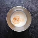 A metal mixing bowl containing cream and vanilla extract sits on a dark textured surface.