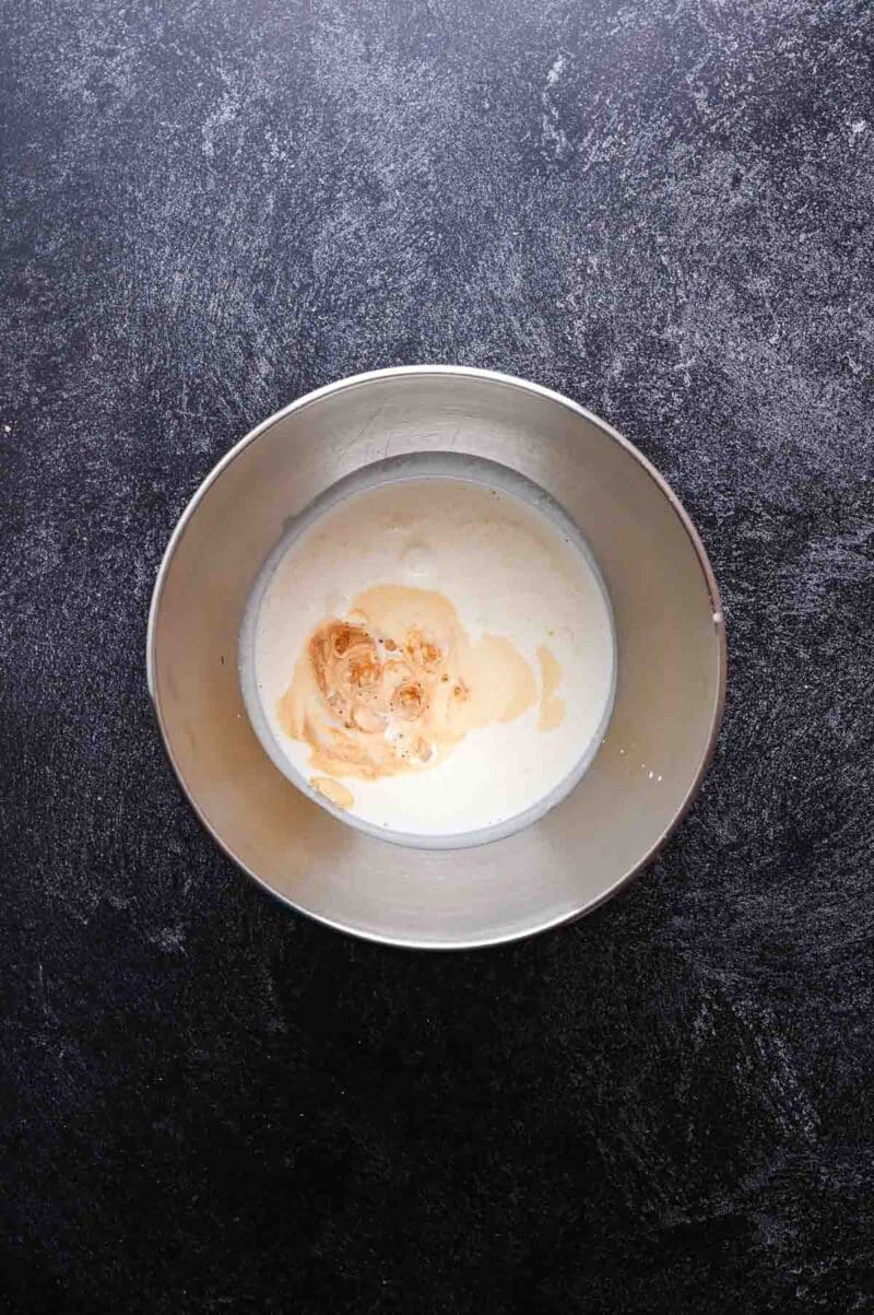 A metal mixing bowl containing cream and vanilla extract sits on a dark textured surface.