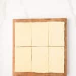 Six rectangular pieces of raw puff pastry are arranged on a wooden cutting board with a handle, set on a white marble surface.