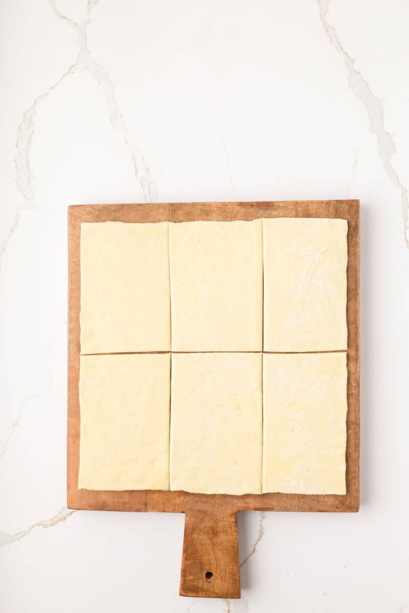 Six rectangular pieces of raw puff pastry are arranged on a wooden cutting board with a handle, set on a white marble surface.