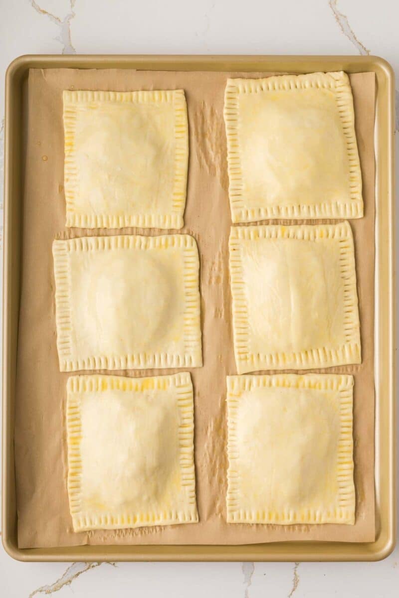 A baking sheet with six unbaked, square hand pies arranged in two columns on parchment paper.