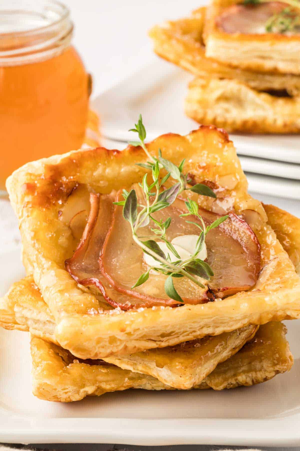Square puff pastry topped with pear slices, herbs, and cheese, stacked on a plate with a jar of honey in the background.
