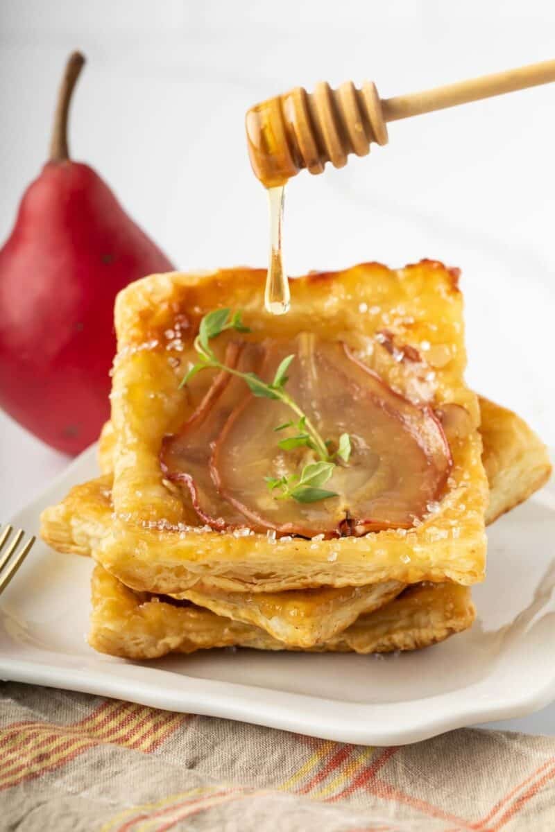 A stack of puff pastry tarts topped with pear slices and herbs, with honey being drizzled from a dipper. A red pear is in the background.