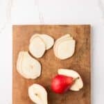 A red pear, some whole and some thinly sliced, is arranged on a wooden cutting board set on a white marble surface.