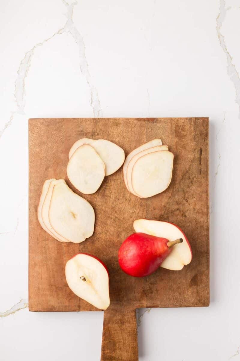 A red pear, some whole and some thinly sliced, is arranged on a wooden cutting board set on a white marble surface.