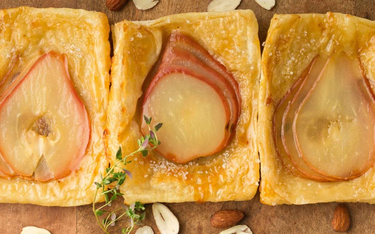 Three square pear tarts with puff pastry and sliced pears are arranged in a row on a wooden surface, garnished with thyme and surrounded by almonds.