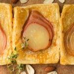 Three square pear tarts with puff pastry and sliced pears are arranged in a row on a wooden surface, garnished with thyme and surrounded by almonds.