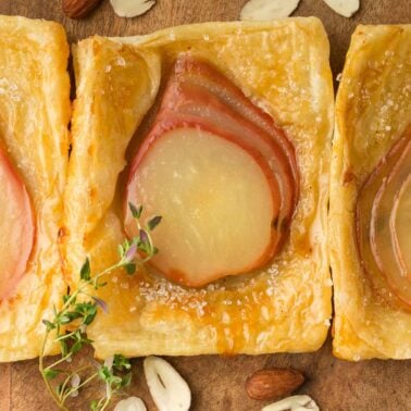 Three square pear tarts with puff pastry and sliced pears are arranged in a row on a wooden surface, garnished with thyme and surrounded by almonds.