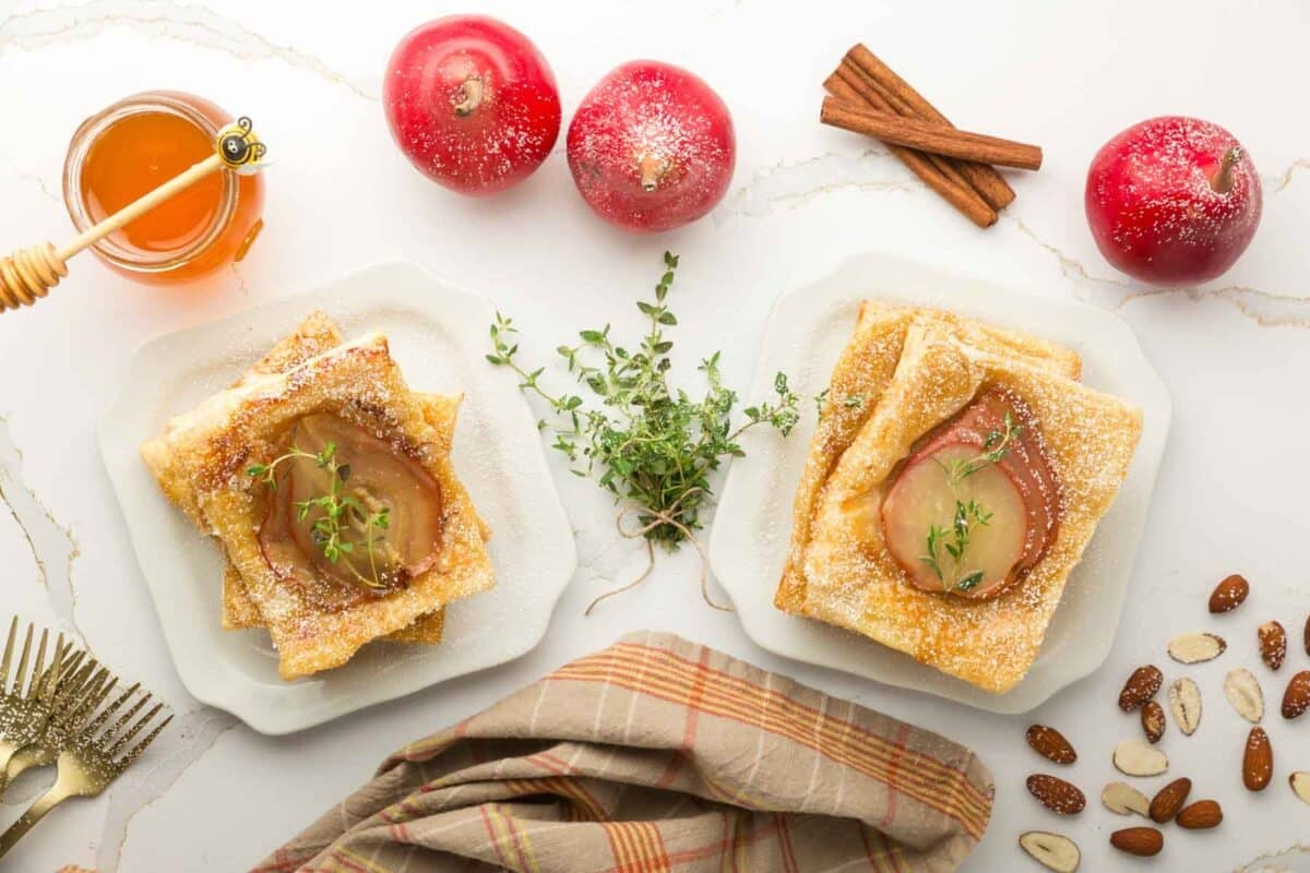 Two plates with puff pastry and baked apple slices, surrounded by apples, honey, cinnamon sticks, nuts, thyme, and a striped cloth on a white marble surface.