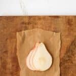Three thin pear slices are arranged on a piece of brown parchment paper on a wooden surface, with a white background above.
