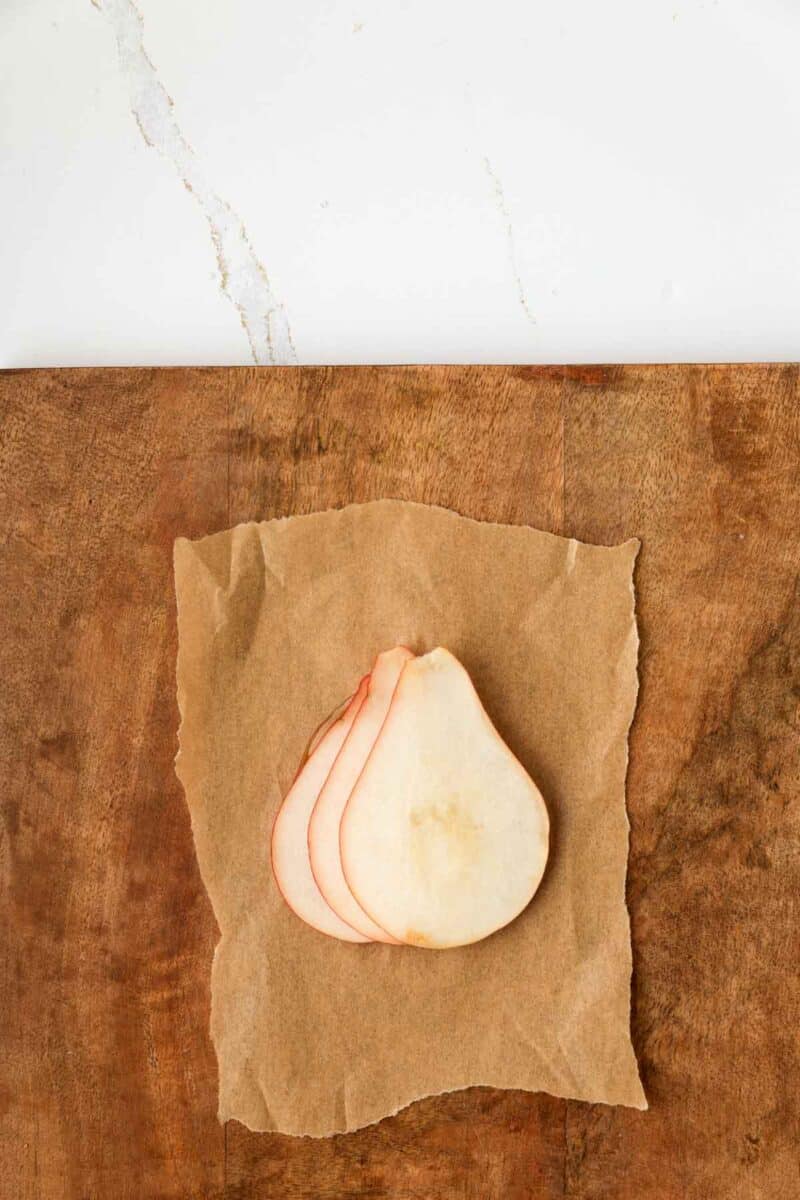 Three thin pear slices are arranged on a piece of brown parchment paper on a wooden surface, with a white background above.