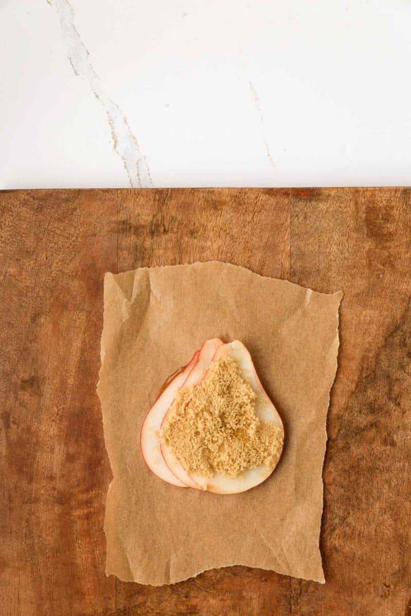 A slice of pear topped with brown sugar sits on a piece of brown parchment paper on a wooden surface, with a white countertop in the background.