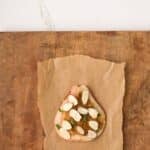 A slice of bread with apple slices, almonds, herbs, and spread sits on brown parchment paper on a wooden surface.