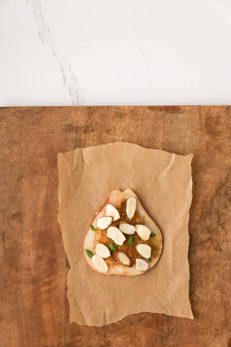 A slice of bread with apple slices, almonds, herbs, and spread sits on brown parchment paper on a wooden surface.