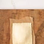 A square, uncooked pastry with crimped edges sits on parchment paper atop a wooden surface.