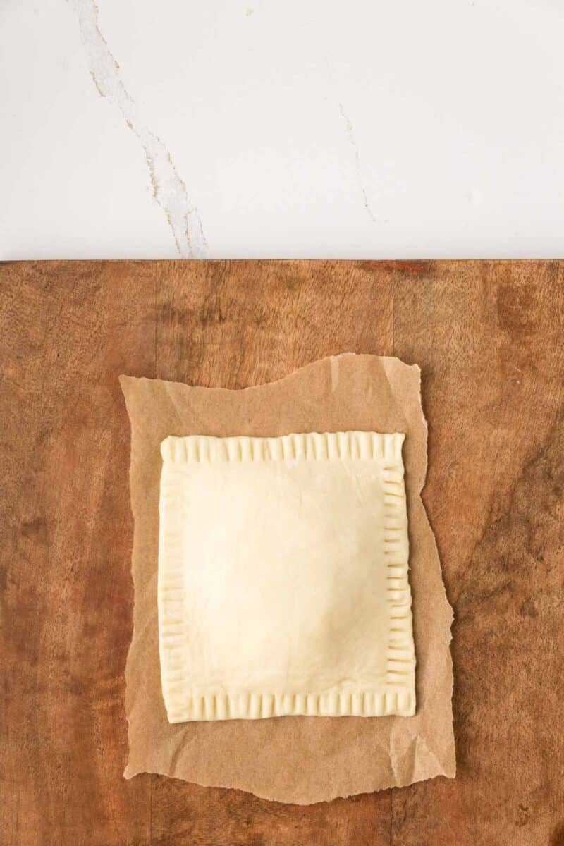 A square, uncooked pastry with crimped edges sits on parchment paper atop a wooden surface.