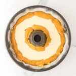 A bundt pan filled with alternating layers of orange batter and white batter, ready to be baked, sits on a white marble surface.
