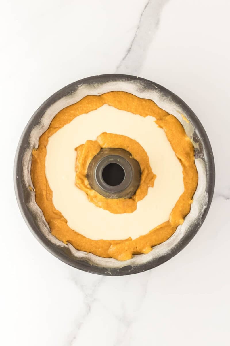 A bundt pan filled with alternating layers of orange batter and white batter, ready to be baked, sits on a white marble surface.