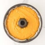 A bundt pan filled with smooth yellow cake batter, ready to be baked, sits on a white marble surface.