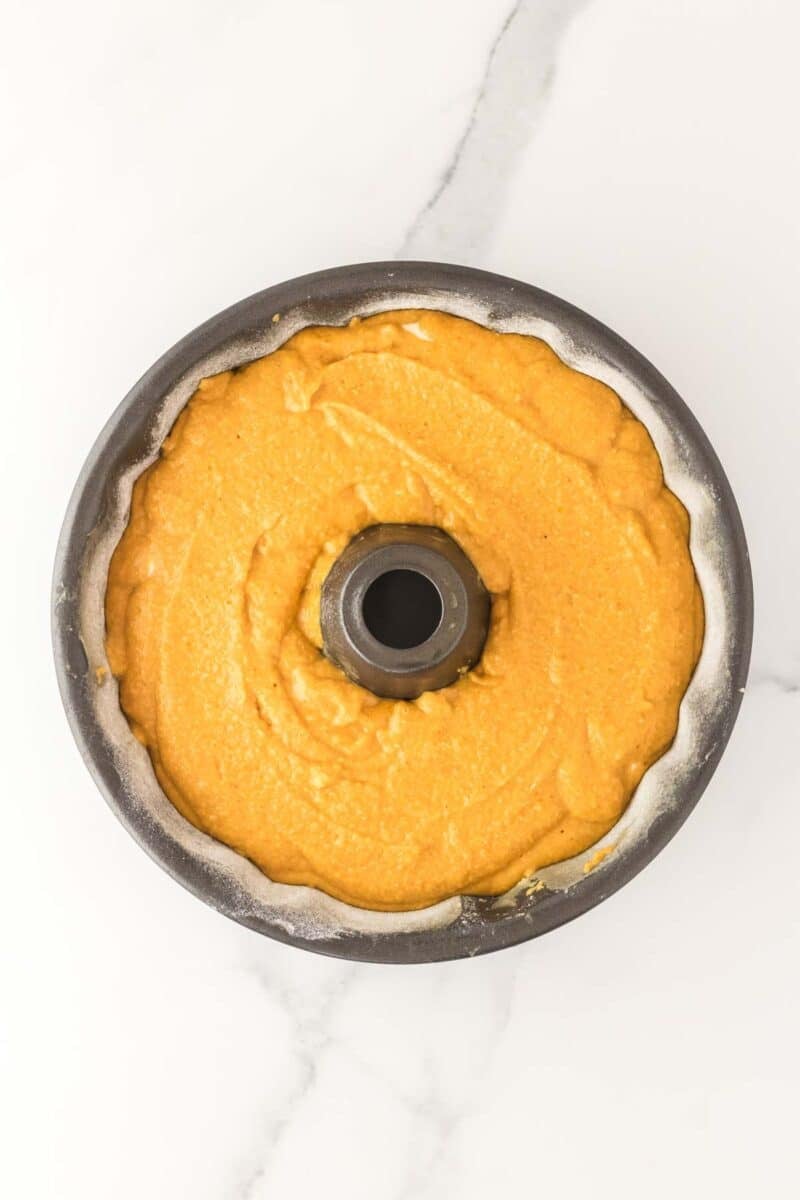 A bundt pan filled with smooth yellow cake batter, ready to be baked, sits on a white marble surface.