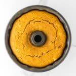 A round, golden-brown Bundt cake with cracks on top, shown from above in a dark Bundt pan on a white marble surface.