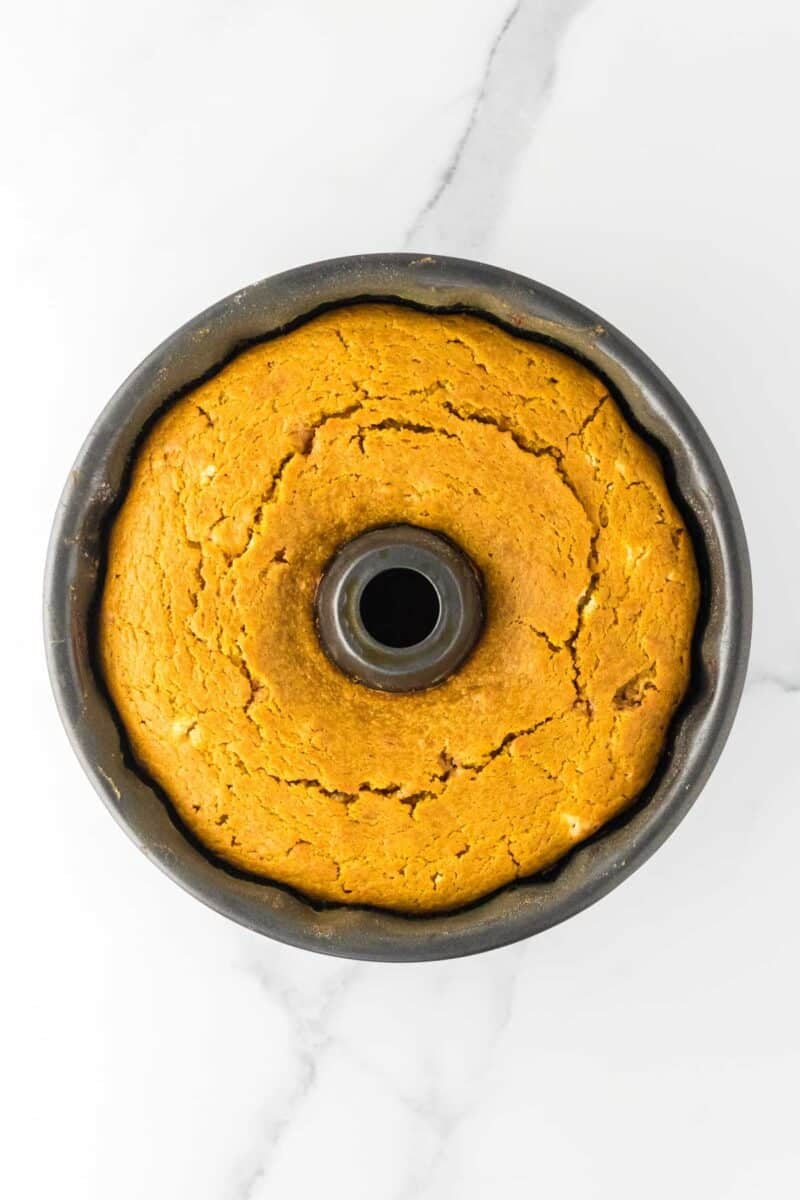 A round, golden-brown Bundt cake with cracks on top, shown from above in a dark Bundt pan on a white marble surface.