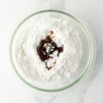 A glass bowl contains powdered sugar with vanilla extract and a small amount of milk on top, sitting on a white marble surface.