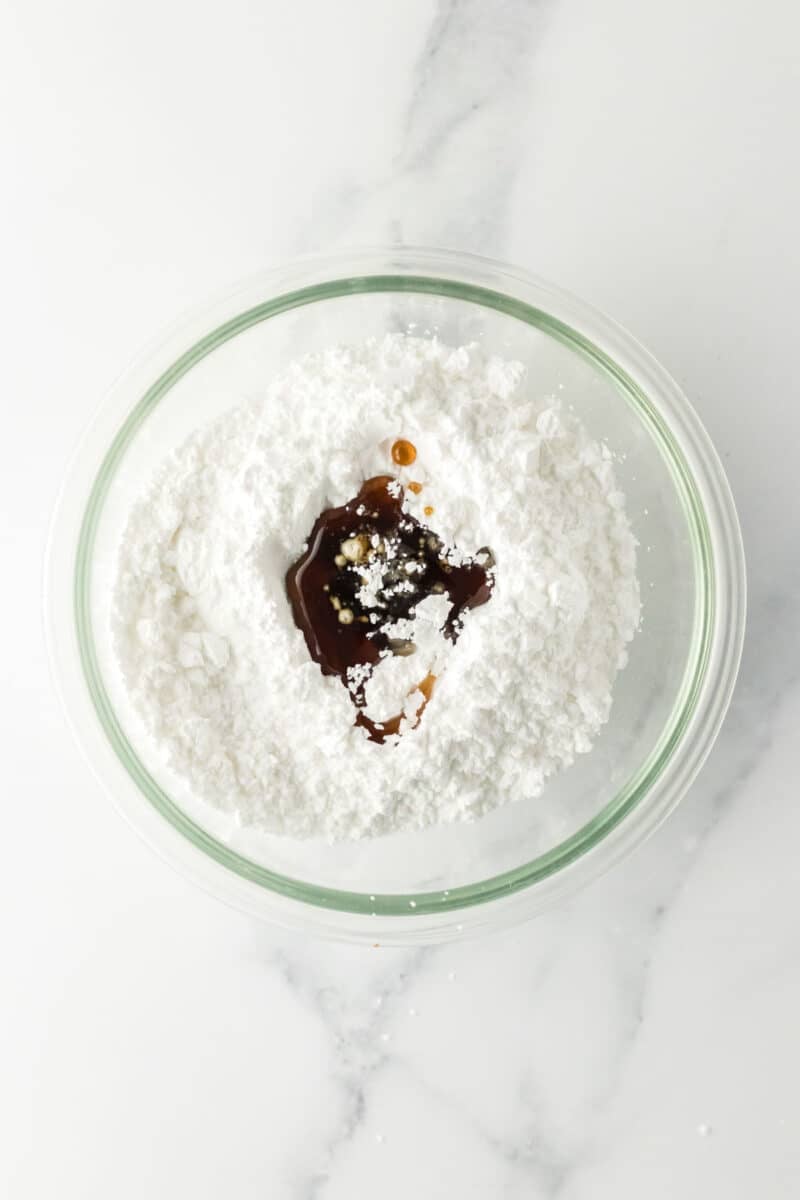 A glass bowl contains powdered sugar with vanilla extract and a small amount of milk on top, sitting on a white marble surface.
