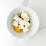 A white bowl containing cream cheese, an egg, powdered sugar, vanilla extract, and sour cream on a marble surface.