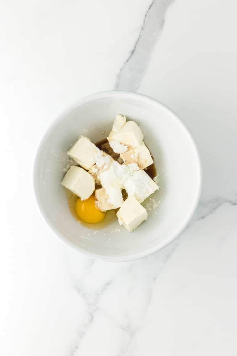 A white bowl containing cream cheese, an egg, powdered sugar, vanilla extract, and sour cream on a marble surface.