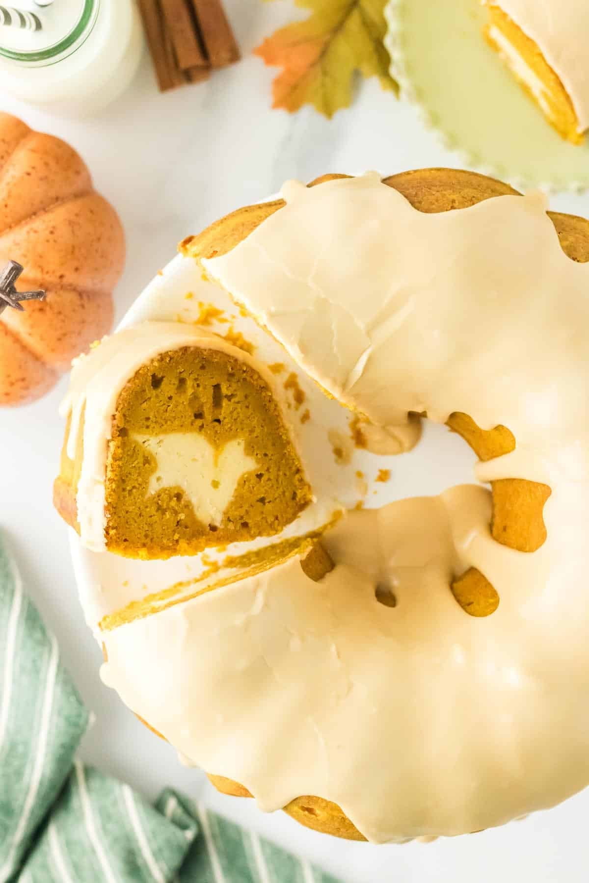 A pumpkin bundt cake with white glaze, sliced to reveal a bat-shaped design inside. Decorative pumpkin, milk, and autumn leaf are nearby.