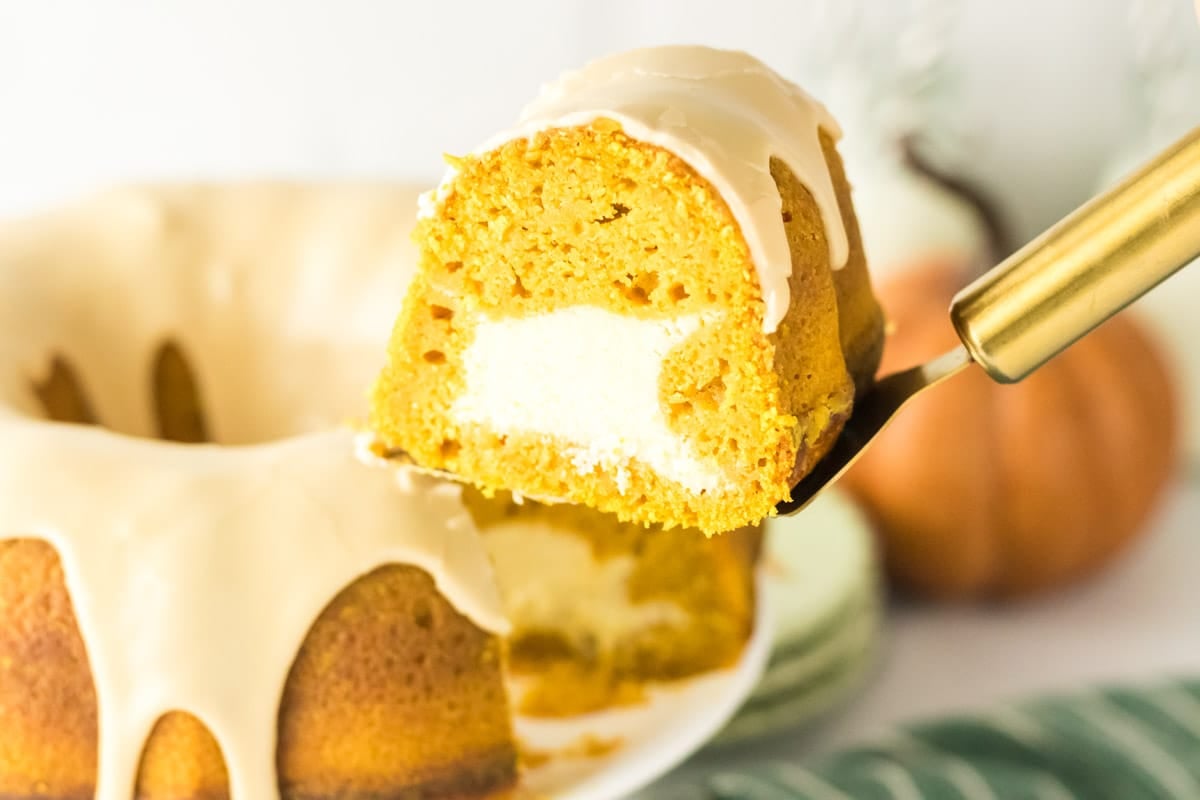A slice of pumpkin bundt cake with a cream cheese filling and glaze is lifted from the whole cake, which sits on a white plate.